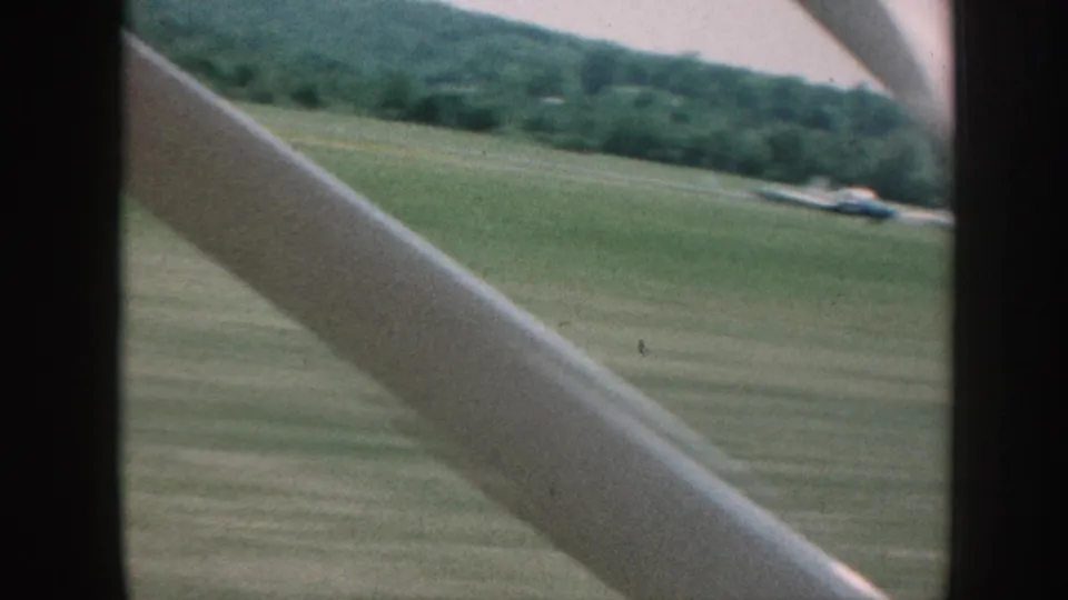 Moving 8mm view over a Nebraska field with a diagonal strut in front, wooded hillside beyond, and a small distant structure or vehicle.