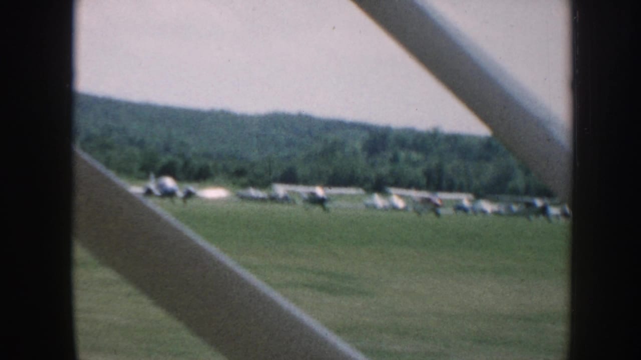 Passenger-side view from a moving aircraft over a Nebraska grass airfield, with several small planes lined up near a tree line.