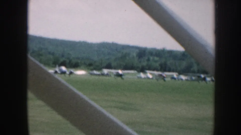 Passenger-side view from a moving aircraft over a Nebraska grass airfield, with several small planes lined up near a tree line.