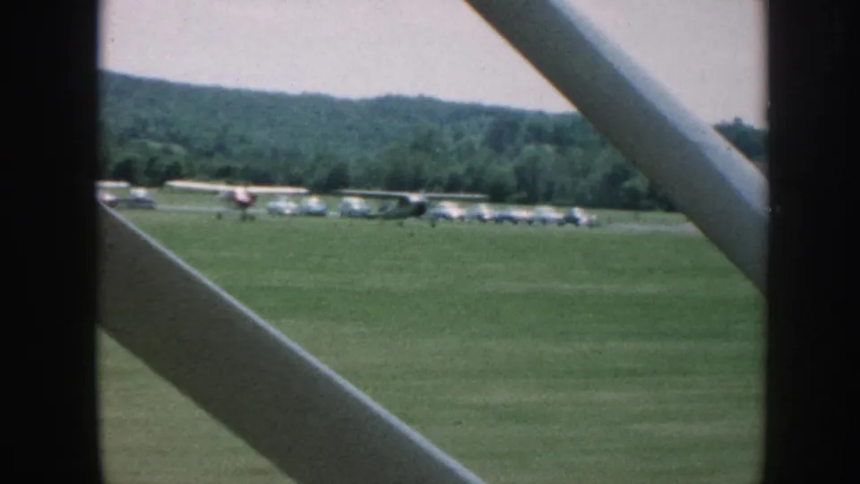 1958 8mm view across a grassy Nebraska airfield, with parked light planes and wooded hills partially obscured by aircraft framing.