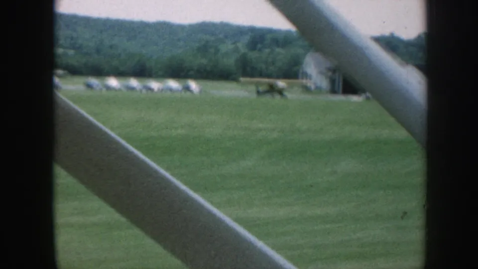 Inside-aircraft view of a Nebraska grass airfield in 1958, showing a light-colored building, parked planes, and foreground struts.