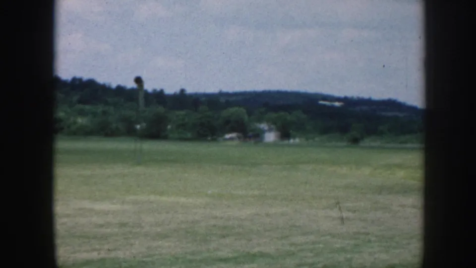 1958 Nebraska home-movie frame showing a grassy airfield through dark vertical window bars, with trees and low hills in the distance.