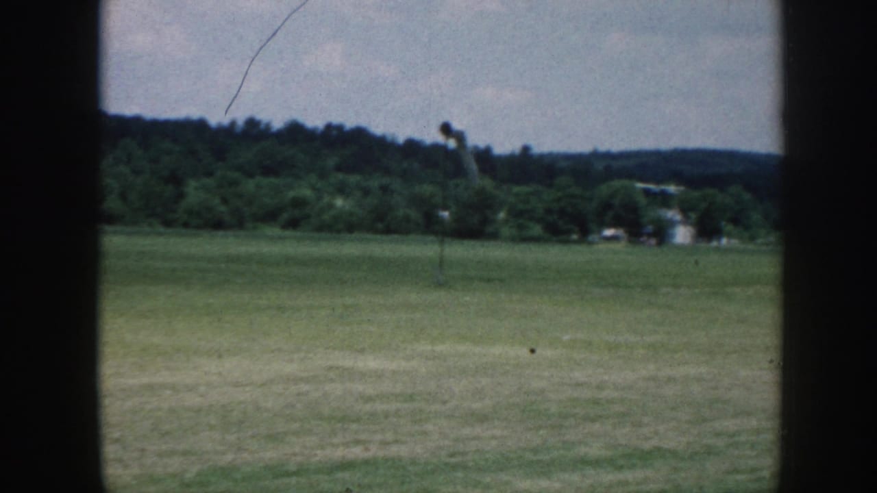 1958 Nebraska frame of an open grass airfield bordered by trees, seen through a dark vertical divider and soft 8mm color.