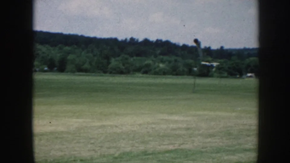 1958 Nebraska home-movie view of a grass airfield, tree line, and a small pole or windsock-like structure near the center.