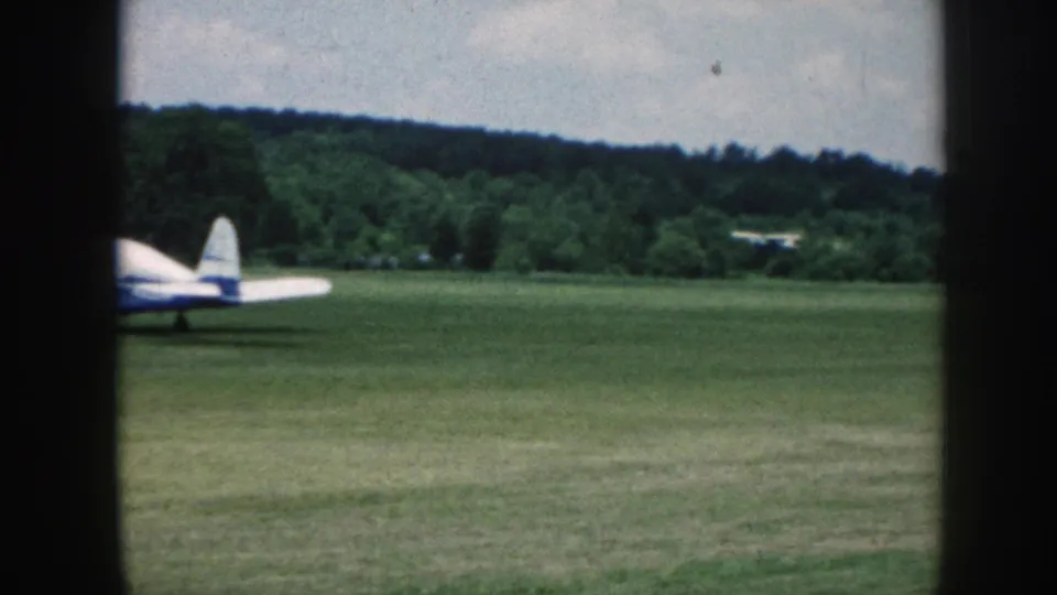 1958 Nebraska frame showing a small white-and-blue airplane on a grass airfield, partly framed by dark window bars.