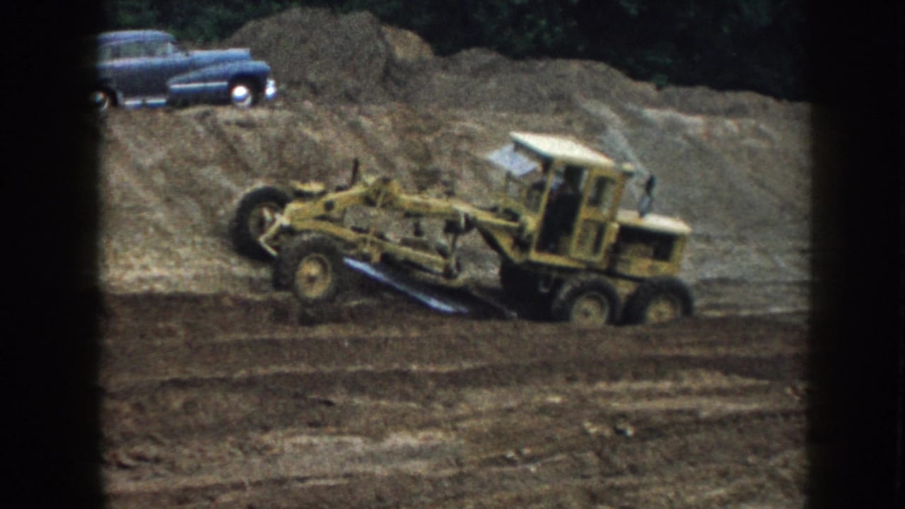 Yellow road grader appears mid-pass on a 1958 Nebraska dirt site, with a blue 1950s sedan parked above the graded earth.