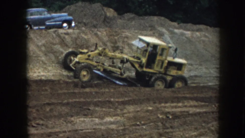 Yellow road grader appears mid-pass on a 1958 Nebraska dirt site, with a blue 1950s sedan parked above the graded earth.