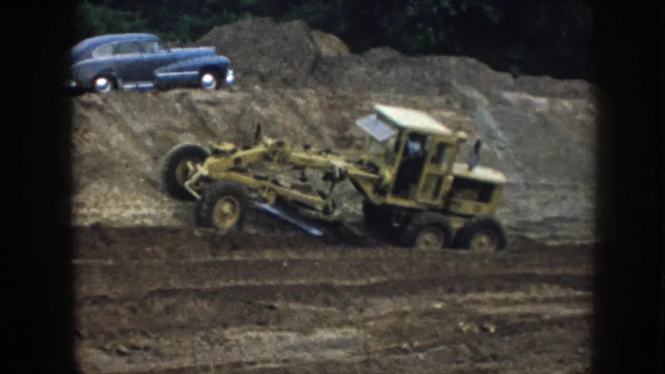 A yellow road grader fills more of the 8mm frame while leveling earth beneath a blue sedan in the Nebraska worksite scene.