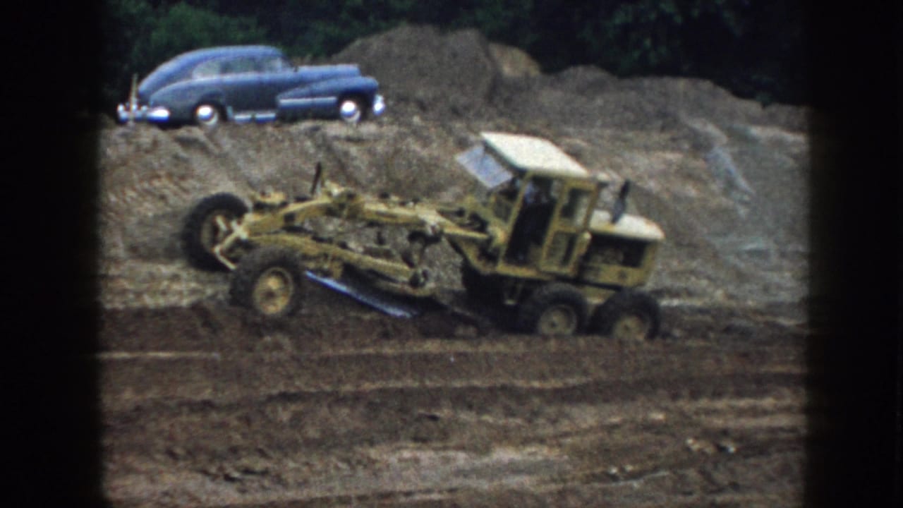 Yellow road grader works across dirt below a blue sedan on an earth mound.