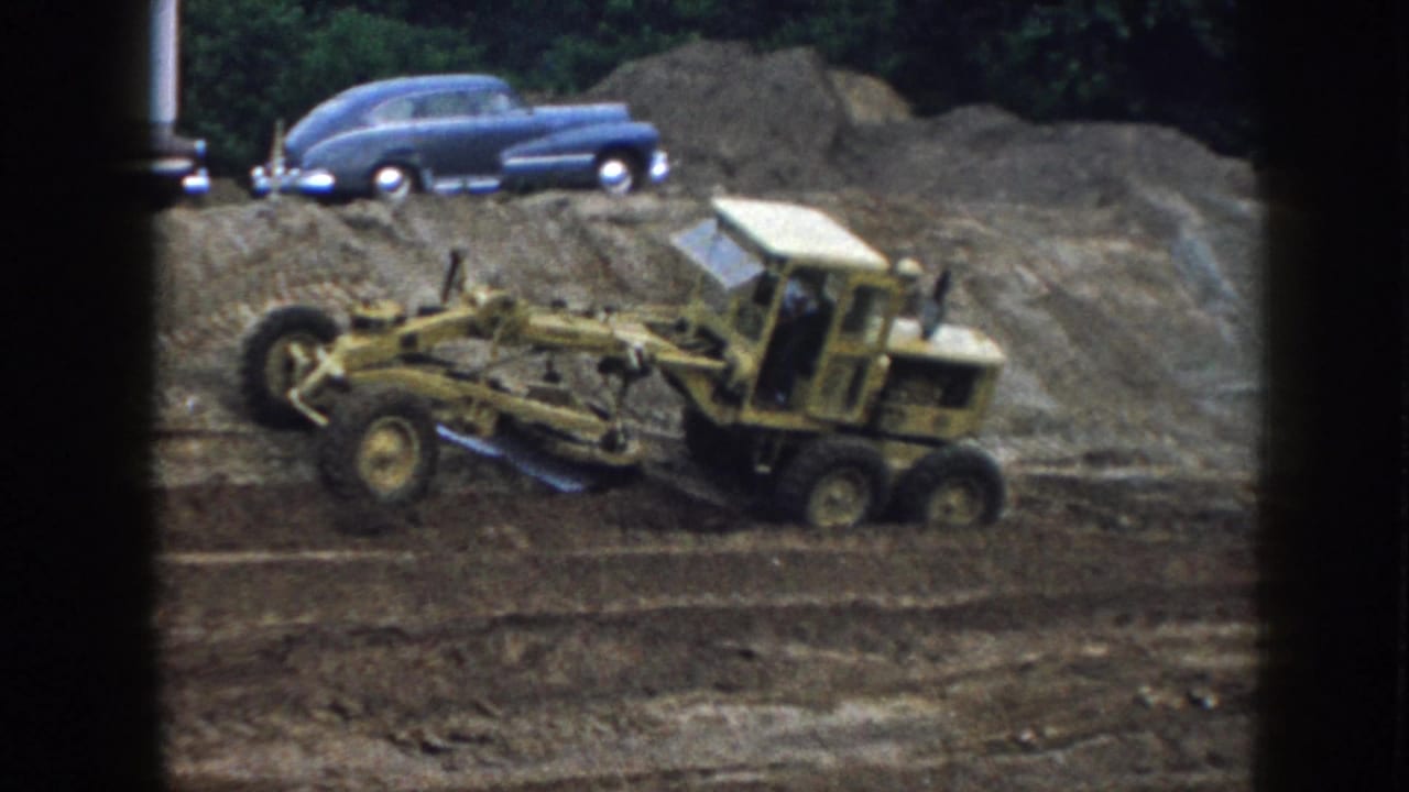 A yellow grader moves across a Nebraska dirt site, with a blue mid-century sedan parked along the raised ground behind it.
