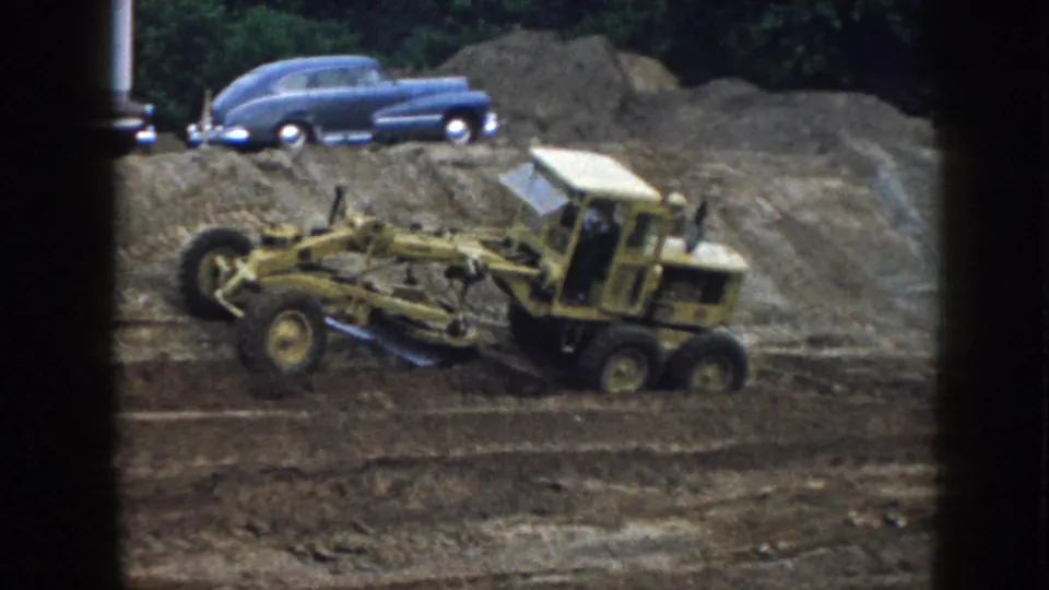 A yellow grader moves across a Nebraska dirt site, with a blue mid-century sedan parked along the raised ground behind it.