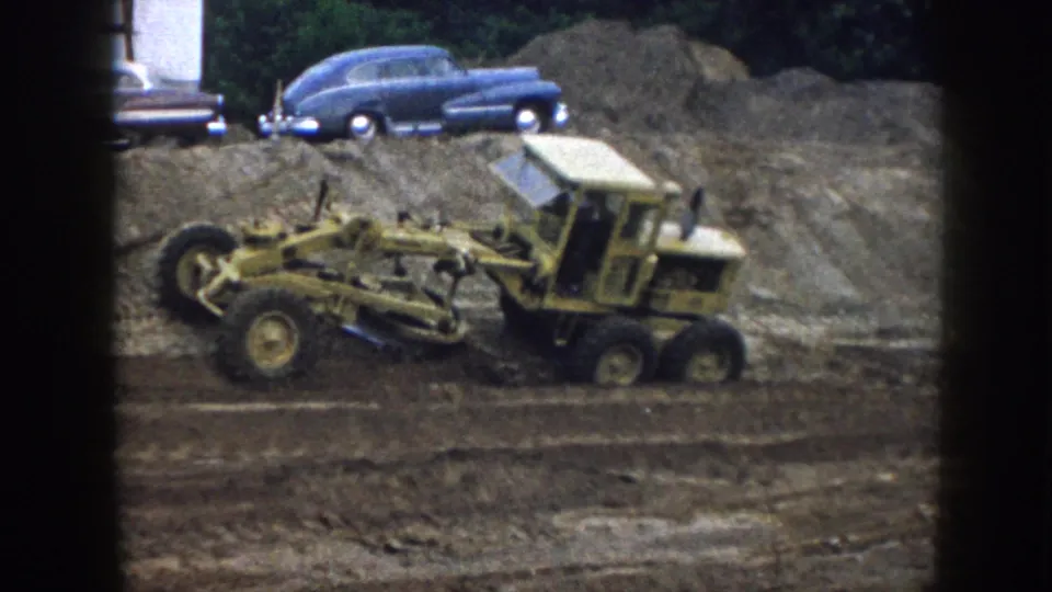 The 8mm frame shows the grader at work on bare ground, with the blue 1950s car and an indistinct vehicle edge in the background.