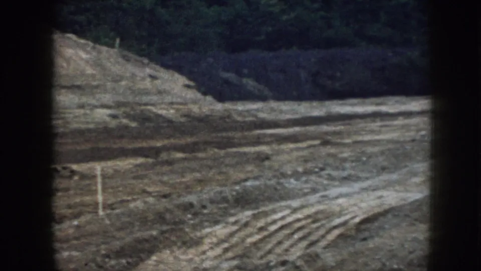 Late-frame view of the Nebraska grading site after the machine has moved out of view, showing dirt tracks, earth banks, and trees.