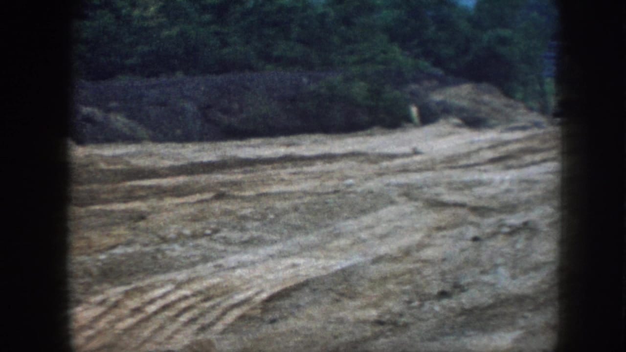 1958 Nebraska 8mm frame showing an open construction lot with churned soil, tire tracks, and trees beyond the graded ground.