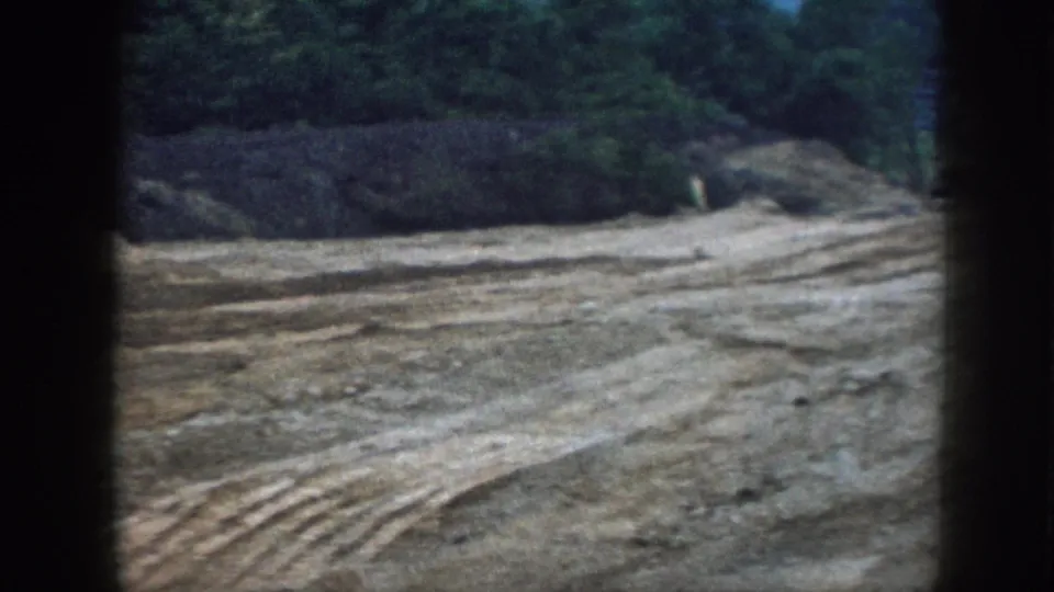 1958 Nebraska 8mm frame showing an open construction lot with churned soil, tire tracks, and trees beyond the graded ground.