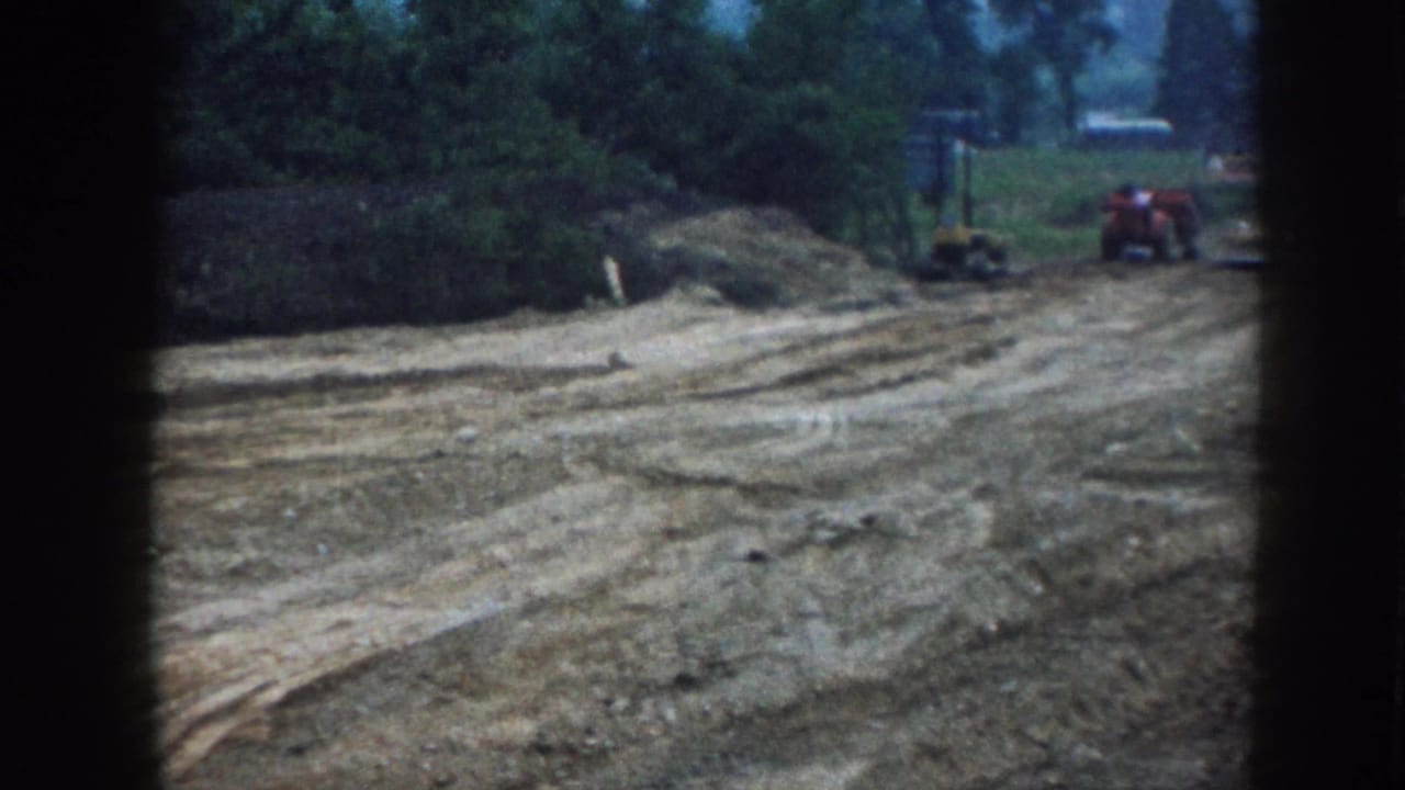 Dirt construction lot with trees, a red tractor, and small equipment in the distance.