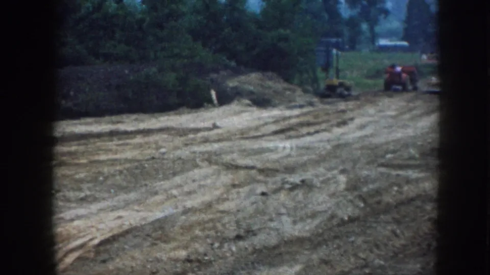 1958 Nebraska construction scene with a red tractor and small equipment beyond a rutted dirt lot near trees.