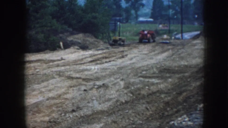 1958 Nebraska 8mm frame showing rutted earth, a red tractor, small construction equipment, and trees around the lot.