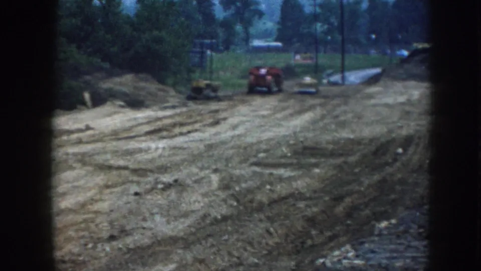 Final 1958 Nebraska frame of the graded lot, with tire-marked soil, a red tractor, small machinery, and trees behind it.