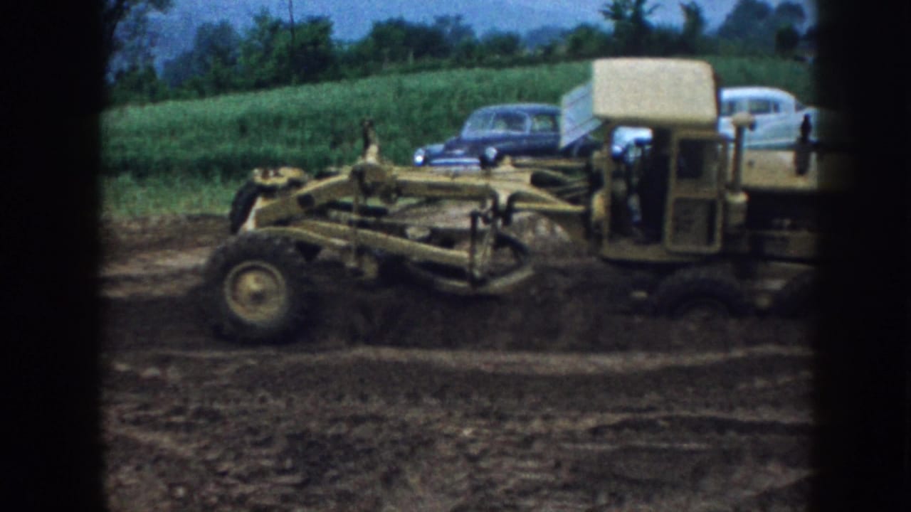 Nebraska 1958 home-movie frame of a yellow road grader leveling dirt beside parked 1950s cars and rural fields.
