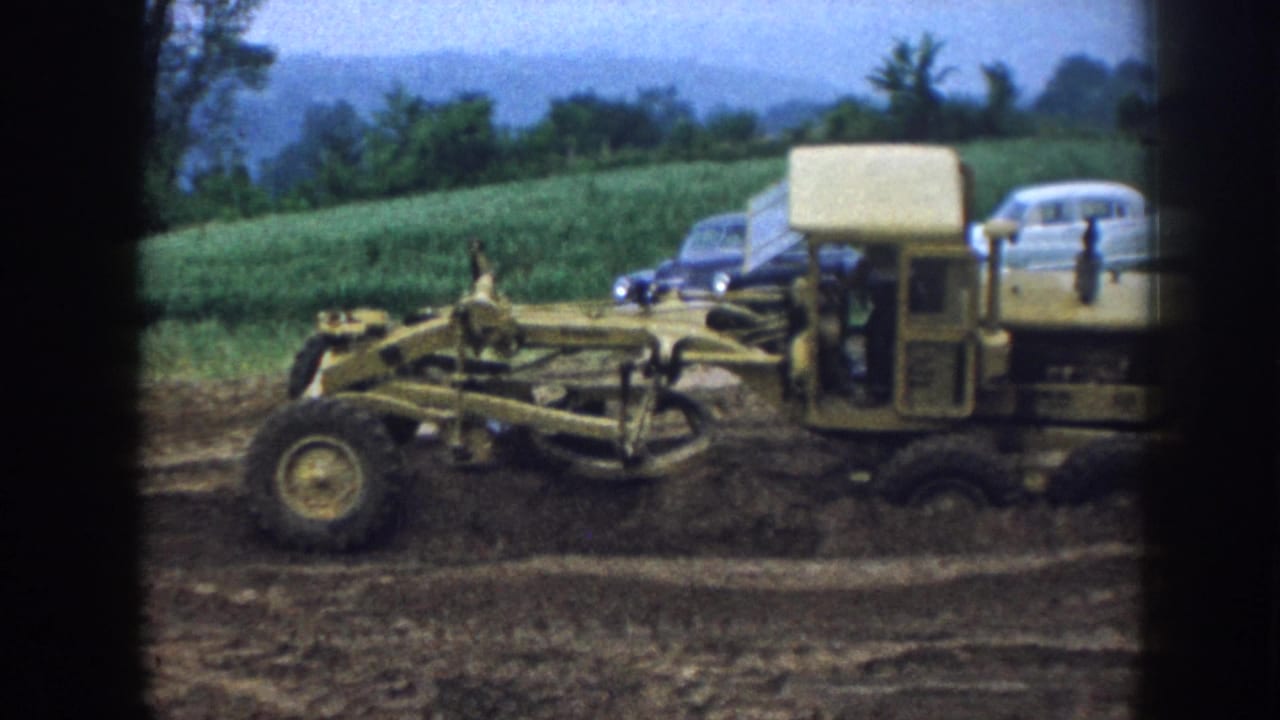 Road grader continues across the dirt with two cars parked near a grassy field in the background.