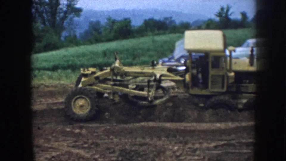 Nebraska 1958 home-movie still of a road grader crossing a muddy work area near a rural field and parked car.