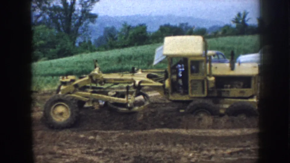 Nebraska 1958 frame with the yellow grader centered on rough dirt, backed by green fields and mid-century automobiles.