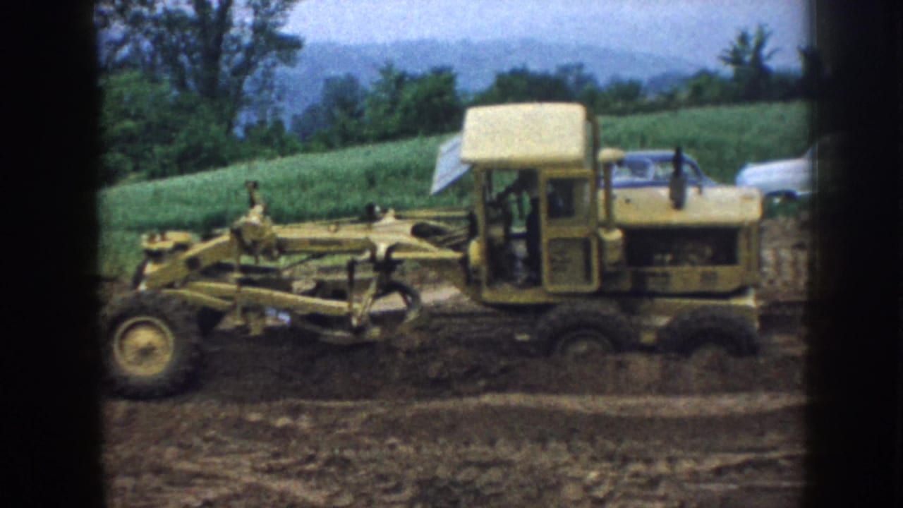 1958 Nebraska 8mm frame showing a yellow grader leveling soil on a rural construction site with green hills and trees in the background.