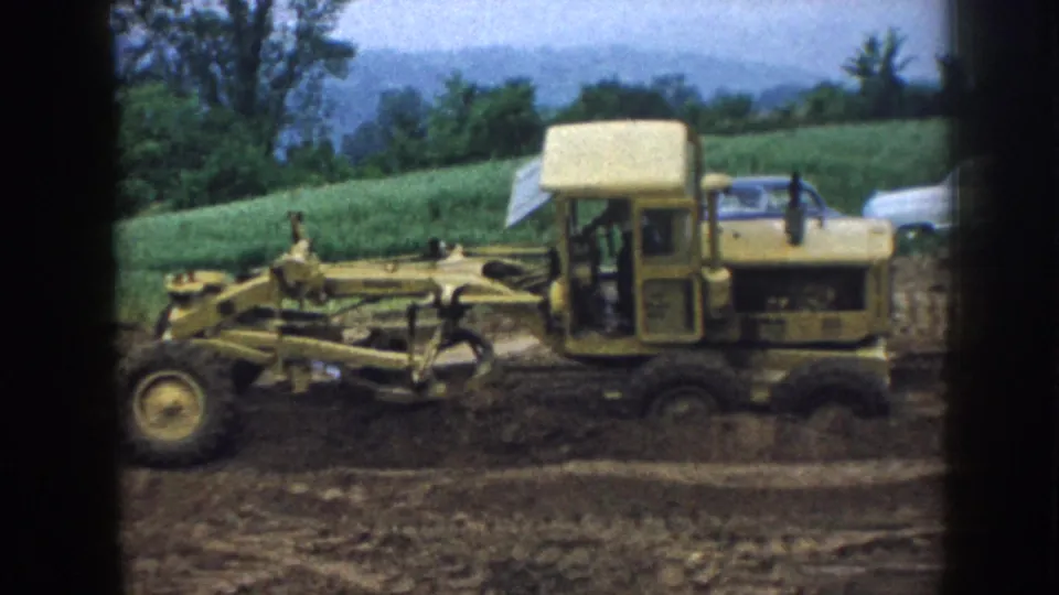 1958 Nebraska 8mm frame showing a yellow grader leveling soil on a rural construction site with green hills and trees in the background.