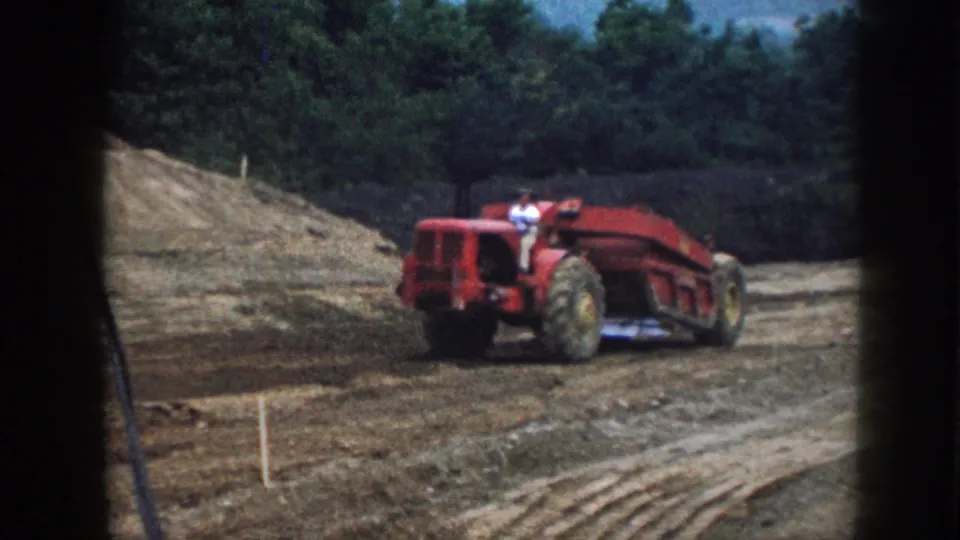 1958 Nebraska 8mm frame showing the rear of a red earthmoving tractor on a muddy rural construction site with trees beyond.