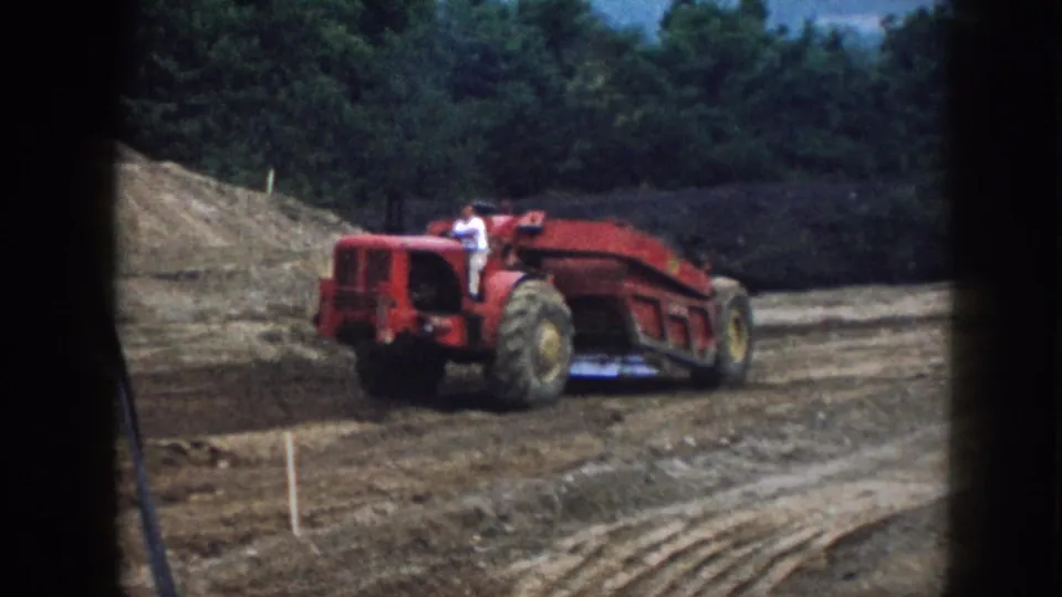 1958 Nebraska 8mm frame showing a red tractor or scraper with an operator on a rural dirt construction site backed by trees.