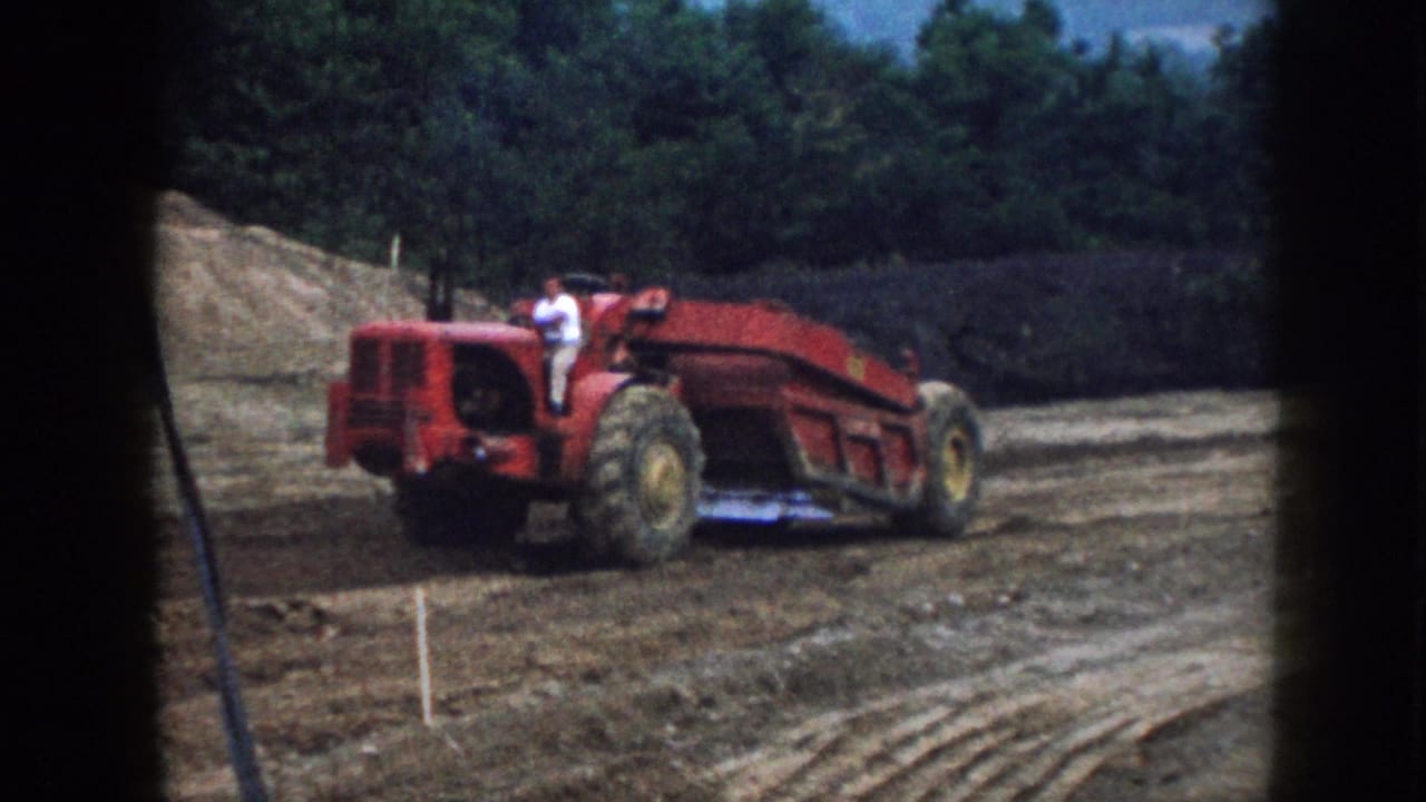 Red tractor with a seated man crosses a dirt field below a wooded hillside.