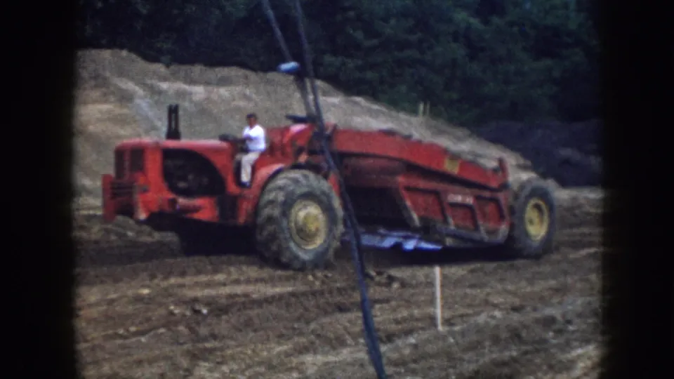 Closer 8mm frame of a man driving a red tractor over rough dirt terrain in Nebraska, with trees and earth banks behind him.