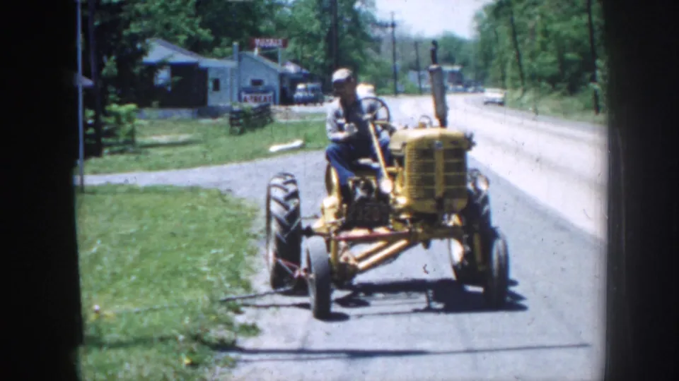 Yellow open-seat tractor travels on a paved rural Nebraska road, with a driver, grass verge, trees, and small roadside buildings visible.