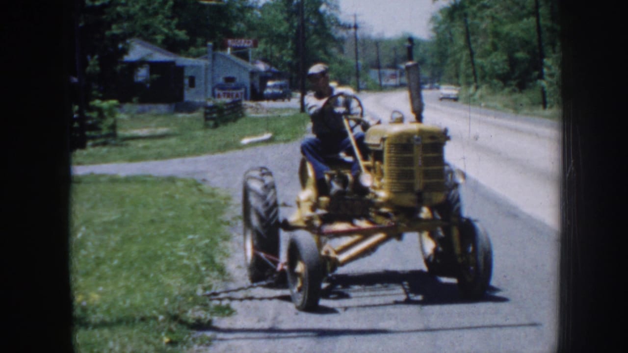 Man drives a yellow tractor along a rural roadside near grass and trees.