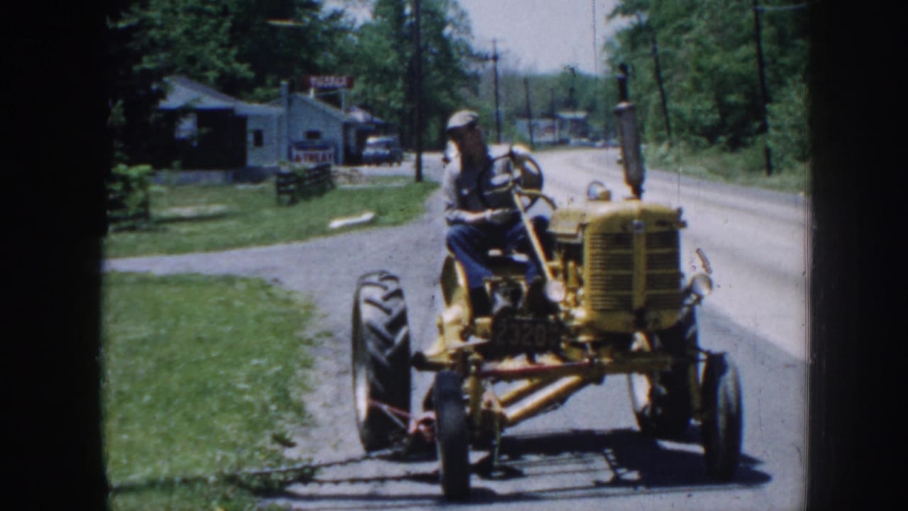 The tractor continues turning on a Nebraska rural road, with the driver seated high and a roadside building, sign, and trees in the background.