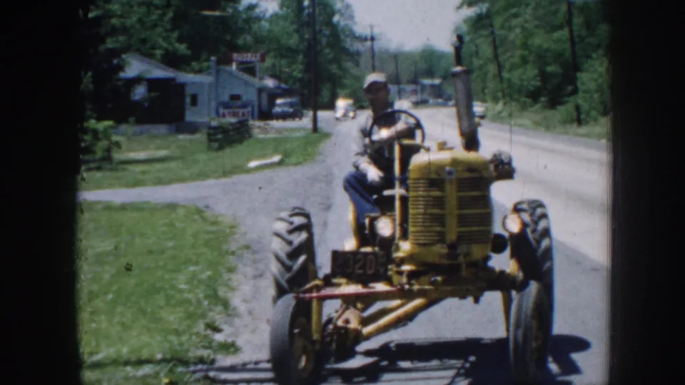 A front-facing view shows the yellow tractor nearing the camera on a 1958 Nebraska road, with a small roadside building and distant cars behind it.