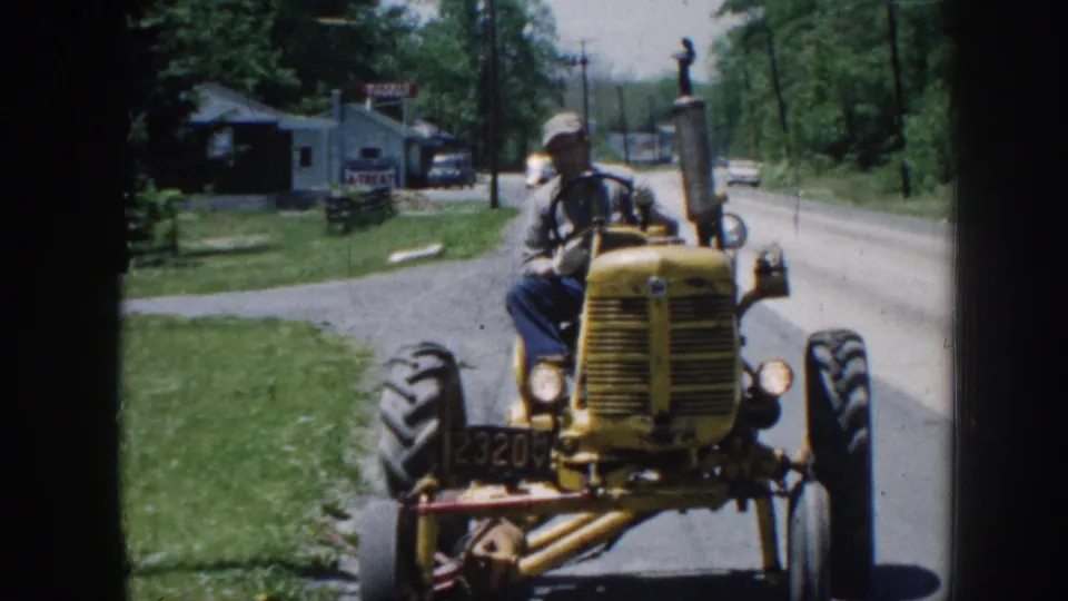 The tractor passes close in a 1958 Nebraska home-movie frame, with the driver in profile and a roadside sign, building, grass, and trees behind him.