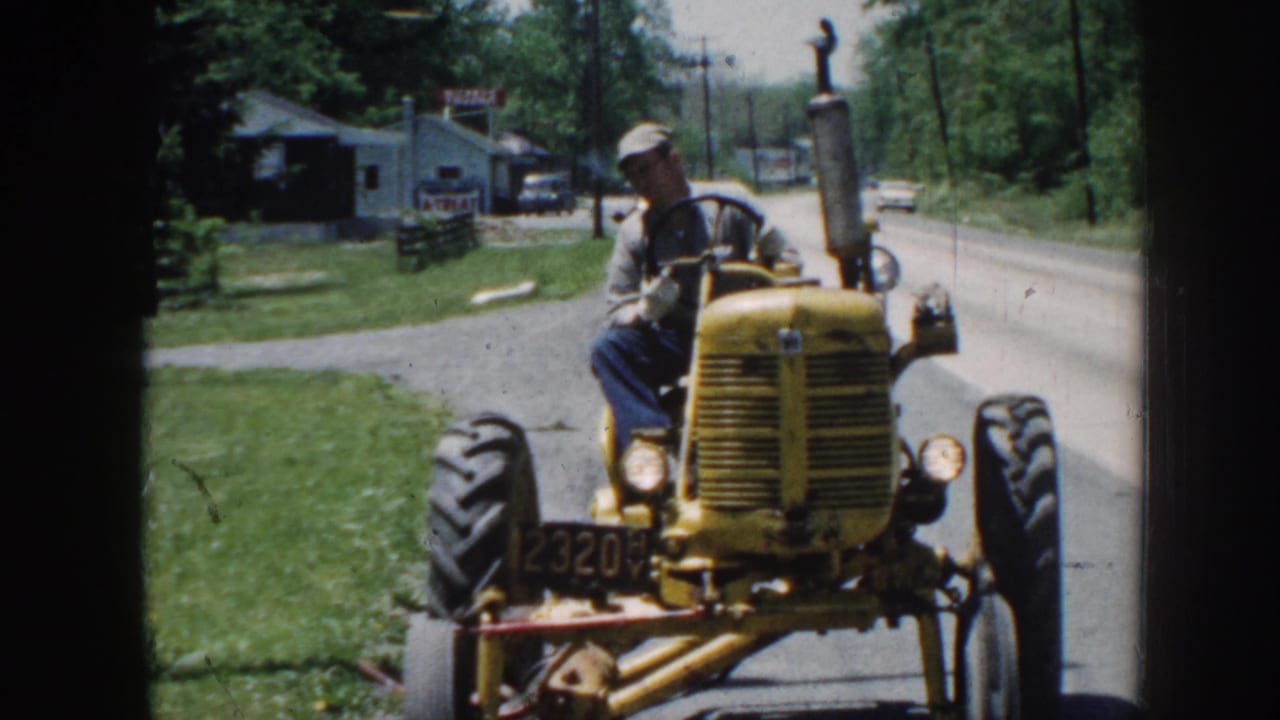 Man in a cap drives a yellow tractor toward the camera on a rural street.