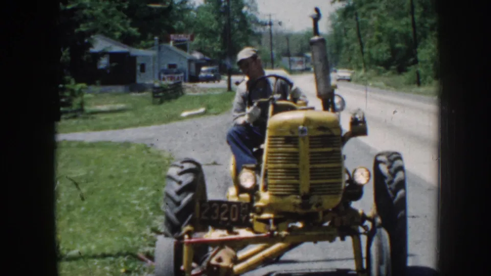 In 1958 Nebraska footage, a man in blue pants drives a yellow tractor toward the camera beside grass, trees, and roadside buildings.