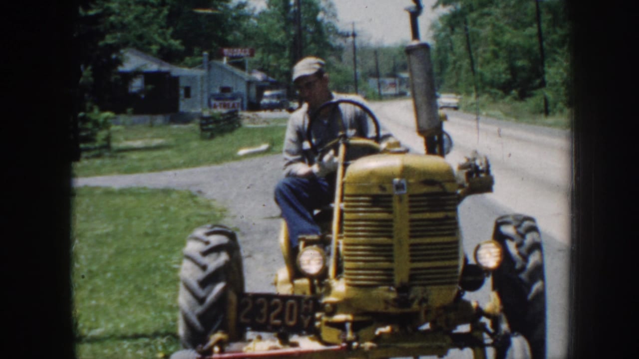 The yellow tractor moves closer on a Nebraska roadside, with the seated driver, large tires, trees, utility poles, and small buildings visible.