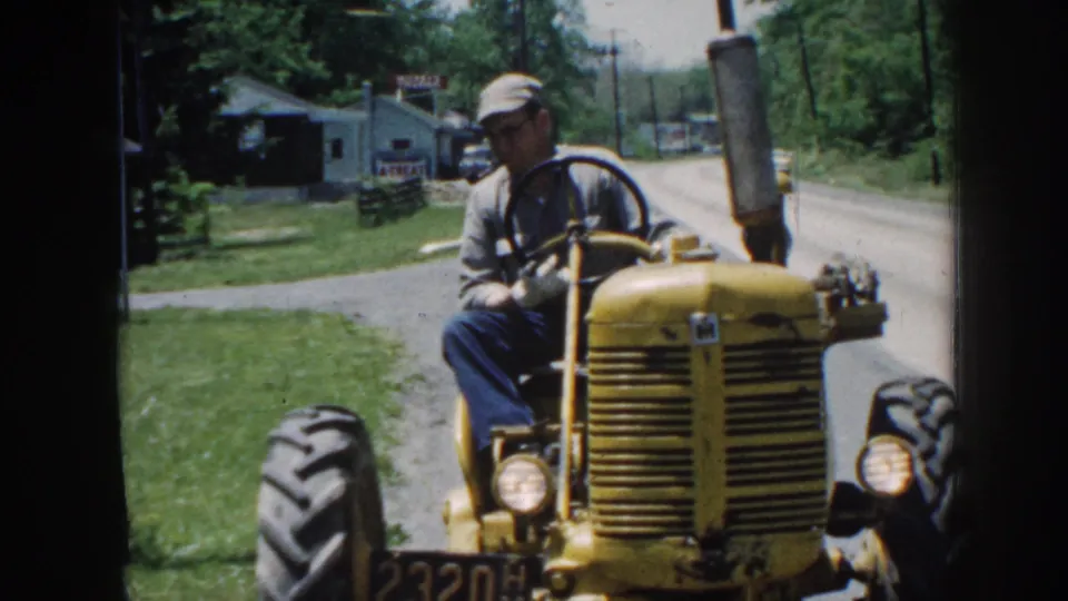 The tractor comes nearer in the 1958 Nebraska home-movie frame, showing the driver, front grille, headlights, grass, trees, and roadside building.