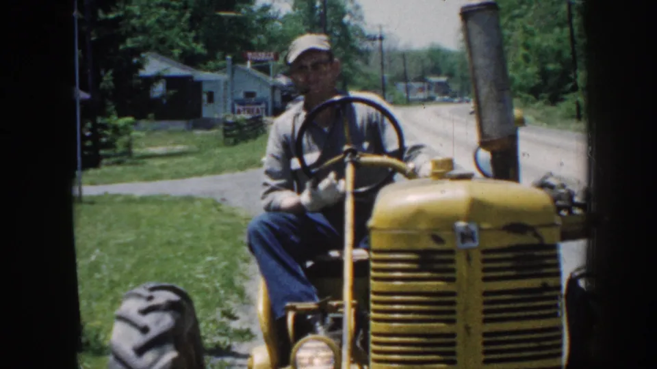 A close 1958 Nebraska frame shows the man at the tractor wheel as the yellow hood, headlight, grass verge, road, trees, and buildings pass behind him.