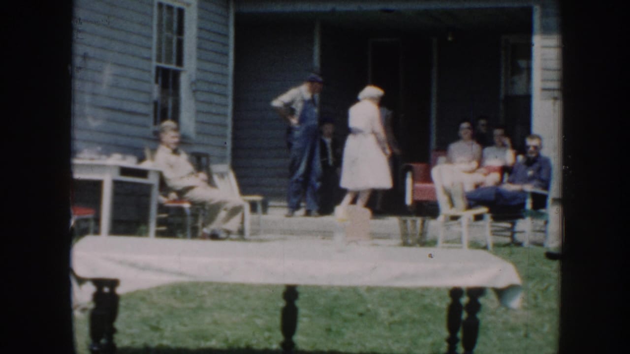 Adults sit and stand outside a clapboard house near a white-covered table on the lawn.