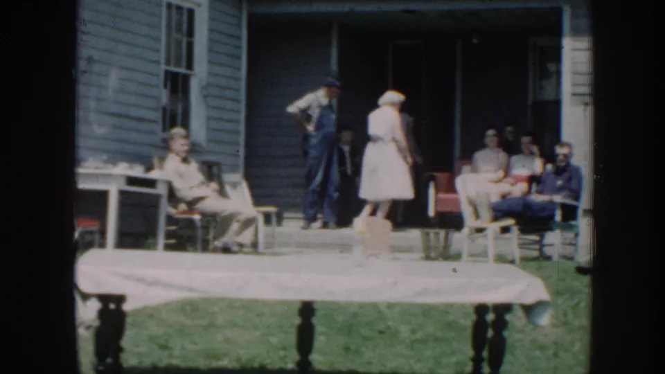 1958 Nebraska home-movie frame showing older adults gathered outside a blue clapboard house, with chairs on the porch and a covered table in front.