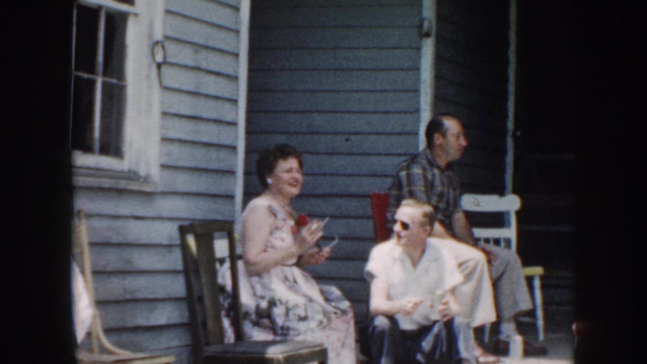 1958 Nebraska porch scene with seated adults in casual dress beside a blue clapboard exterior and a man standing nearby.