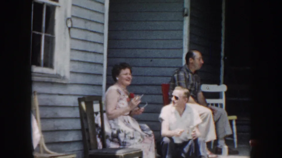 1958 Nebraska porch scene with seated adults in casual dress beside a blue clapboard exterior and a man standing nearby.