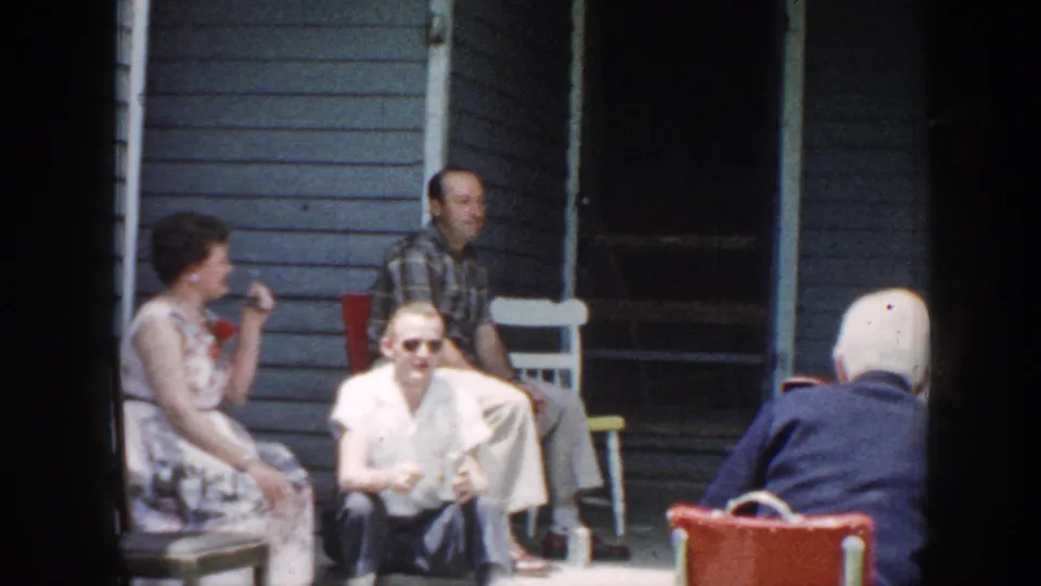 1958 Nebraska home-movie frame showing older adults seated near a shaded porch doorway and clapboard wall during an outdoor visit.