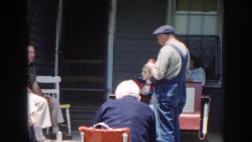 1958 Nebraska frame showing a man in blue overalls standing on the porch area while older adults sit in chairs nearby.