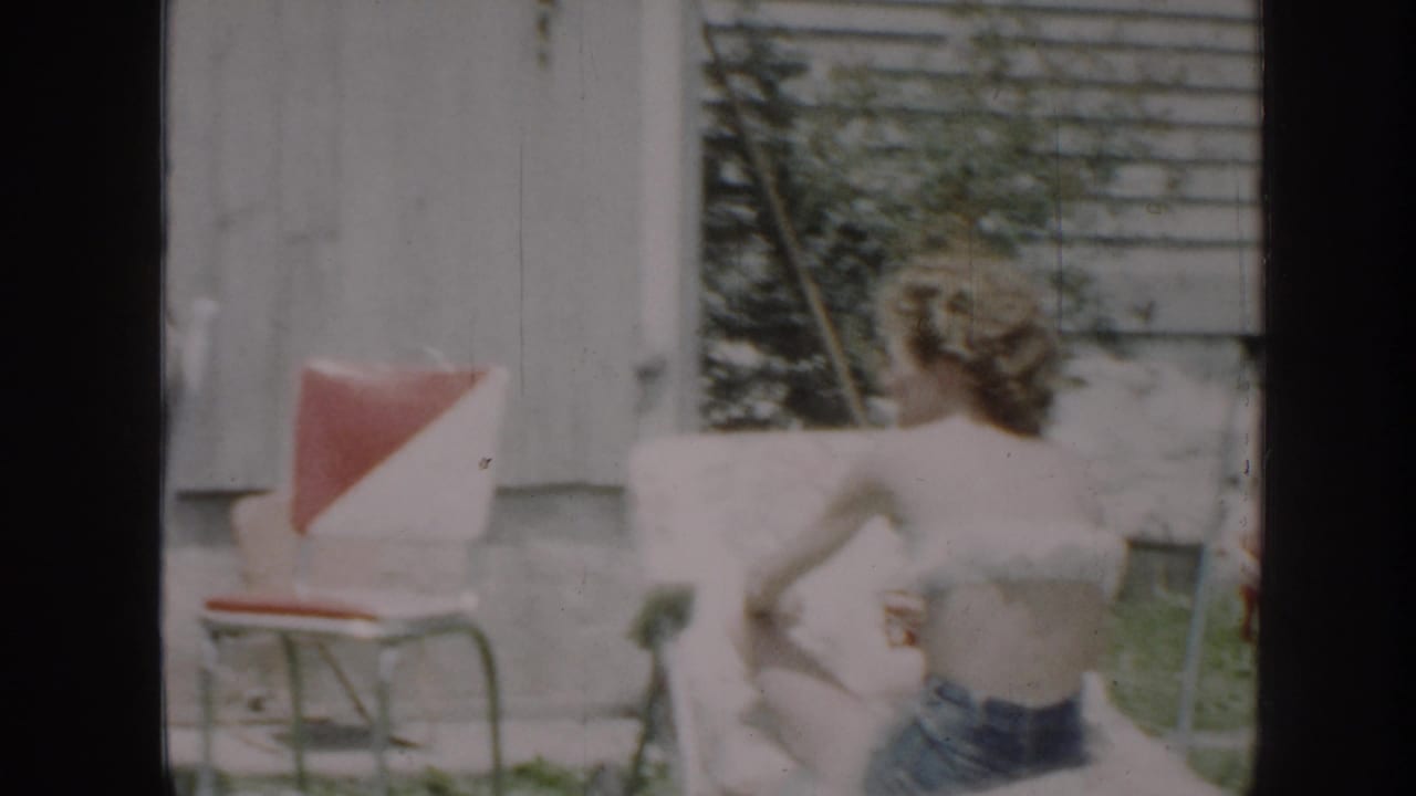 1958 Nebraska home-movie frame showing a woman reclining in a backyard lawn chair beside a red-and-white outdoor chair.